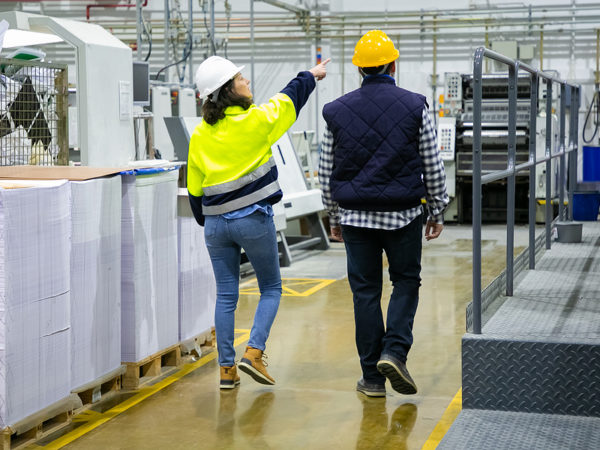 Back view of engineer and inspector pointing fingers at machines in workshop. Man and woman in hardhats and uniform walking on plant floor. Labor or occupation concept