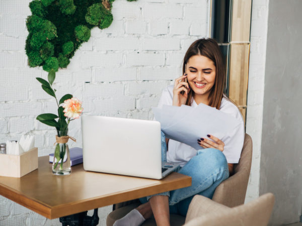 young freelancer girl talking on the phone, holding a notebook, laptop on the table, Beautiful Young Freelancer Woman Using Laptop Computer Sitting At Cafe Table. Happy Smiling Girl Working Online Or Studying And Learning While Using Notebook. Freelance W